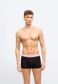 Νέος άνδρας με σγουρά μαλλιά, γυμνός από τη μέση και πάνω, φοράει μαύρα μποξεράκια με ροζ ζώνη Tommy Hilfiger, στέκεται μπροστά σε απλό λευκό φόντο.