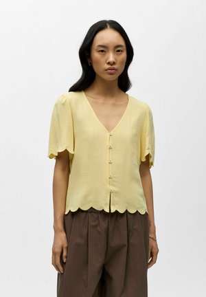 Kvinne iført en lysegul bluse med bølget kant, korte ermer og knapper foran, kombinert med løse brune bukser.