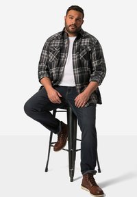 Ternet sort og hvid flannel skjorte over en hvid t-shirt, parret med mørke jeans og brune snøre støvler, siddende på en barstol.