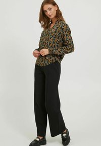 Schwarze Bluse mit langen Ärmeln und gerafftem Bund, verziert mit orangefarbenen und blauen Blumen auf dunklem Hintergrund. Kombiniert mit weiten schwarzen Hosen und klobigen Schuhen.
