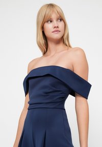 Morska haljina s otvorenim ramenima koja se odlikuje strukturiranim, nabranim dizajnom, mekim tekstilom i širokim rukavima, stvarajući elegantnu siluetu.
