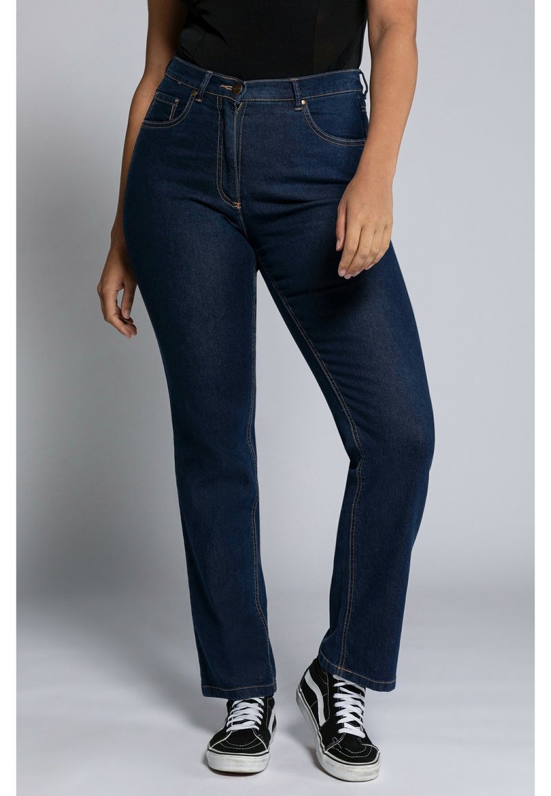 Jean en denim bleu foncé avec une coupe droite, taille haute, fermeture par bouton supérieur et détails de couture subtils ; accompagnés de baskets noires.