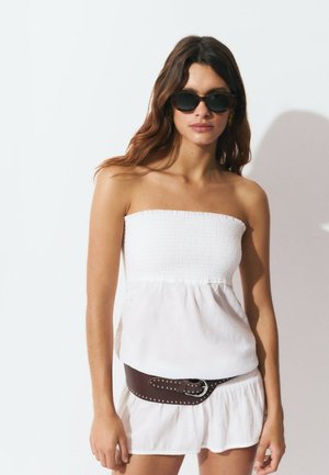Top blanco sin mangas con textura fruncida, combinado con una falda blanca fluida. Cinturón marrón con tachuelas alrededor de la cintura. Los accesorios incluyen gafas de sol negras.