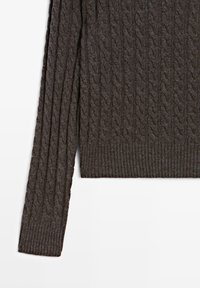 Manche et torse d'un pull en maille gris foncé avec motif torsadé et poignets et ourlet côtelés sur fond blanc.