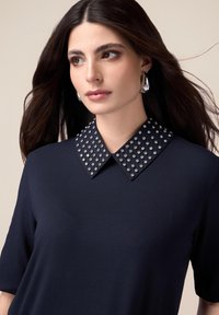 Haut bleu marine avec un col clouté en tissu ; texture lisse ; manches courtes ; présente des clous argentés sur le col.