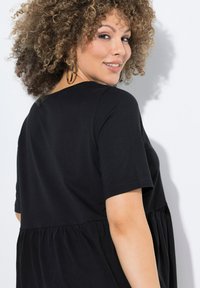 Černý top s krátkým rukávem, přiléhavým korzetem a volně střiženým, nařaseným peplum. Měkká látka, kulatý výstřih a decentní prošívací detaily.