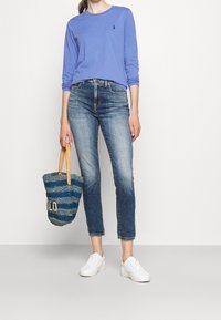 Chemise à manches longues bleu clair avec logo, jean droit délavé moyen, baskets blanches et sac en osier bleu avec les lettres "LO".