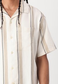 Chemise à manches courtes avec boutons de couleur crème, ornée de rayures verticales beiges et noires, dotée d'une poche poitrine et d'un design texturé.