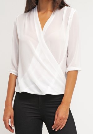 Femme portant un chemisier blanc transparent à devant croisé avec des manches trois-quarts et un pantalon noir, posant avec une main près de la hanche.