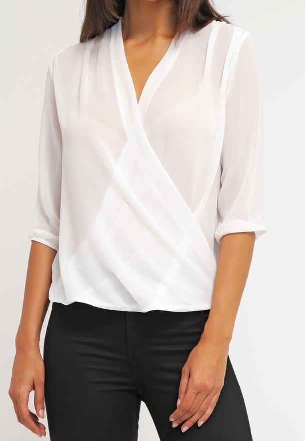 Femme portant un chemisier blanc transparent à devant croisé avec des manches trois-quarts et un pantalon noir, posant avec une main près de la hanche.