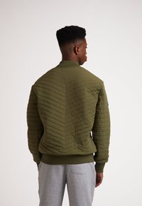 Giacca verde oliva con motivo trapuntato, colletto, polsini e orlo a coste. Presenta un design aderente e una trama liscia.
