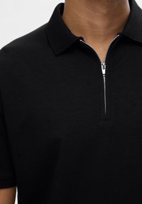 Svart pikétröja tillverkad av mjukt tyg med en kort krage som har dragkedja. Har en subtil textur på kragen och en ren, enkel design.