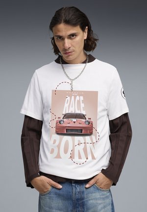 Mann mit schulterlangem Haar trägt weißes T-Shirt mit rosa Rennwagen und dem Schriftzug "RACE BORN", kombiniert mit braunen Langarmshirt und Jeans.
