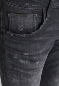 Vaqueros de mezclilla de lavado oscuro con una textura desgastada, con un bolsillo trasero que incluye una etiqueta negra. Presentan detalles sutiles de decoloración y desgaste.