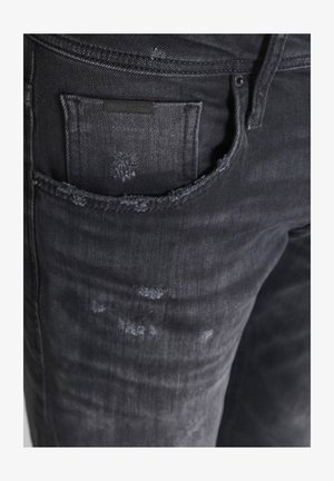 Tamne traperice tamnog pranja s istrošenom teksturom, s stražnjim džepom koji ima crnu oznaku. Prisutni su suptilni efekti blijeđenja i habanja.