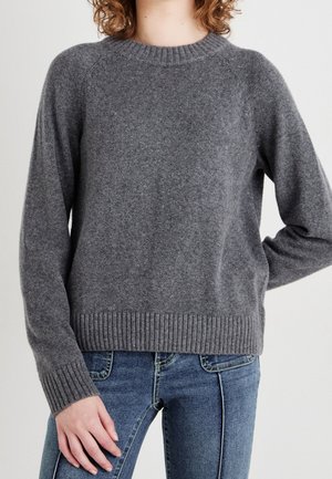 Person iført en grå strikket sweater med ribstrikkede manchetter og kant, kombineret med blå denimjeans.