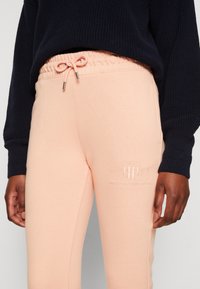 Persoon in lichtroze sweatpants met trekkoord en geborduurd logo, en een donkerblauwe gebreide trui, met ontspannen handen langs de zijde.