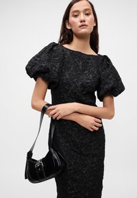Črna cvetna čipkasta obleka s napihnjenimi rokavi, prileganja silhuete in teksturirano blago. V kombinaciji z majhno črno torbico z detajlom s sponko.