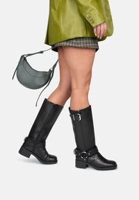 Zwarte leren kniehoge laarzen met een chunky hak en zilveren hardware. Gepaard met een groene leren blazer en een kleine grijze handtas.
