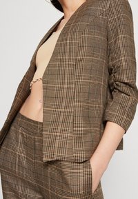 Blazer à carreaux marron avec une coupe décontractée, doté d'un col cranté, de manches trois-quarts et de détails subtils sur les poches.