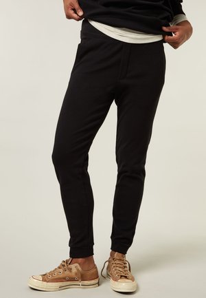 Pantaloni felpati neri con una vestibilità slim, morbido materiale in cotone, vita elastica e tasche laterali, abbinati a sneakers in tela marroni.