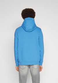 Felpa blu con tasca a marsupio e cappuccio con coulisse, realizzata in tessuto morbido. Indossata con jeans grigio chiaro.