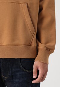 Sudadera marrón con un bolsillo canguro, puños y dobladillo acanalados, hecha de tejido texturizado. Llevada sobre jeans de mezclilla oscuros.