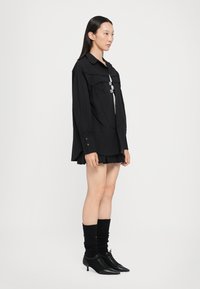 Camisa negra de gran tamaño con un cuello amplio, bolsillos frontales y detalles de botones, combinada con una falda plisada y botas negras de tobillo.