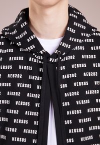 Nära bild av en person som bär en svart hoodie med vit "VERSUS" textmönster över en vit t-shirt, som visar halsen och övre delen av bröstet.