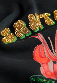 Czarna tkanina z kwiatowym wzorem w pomarańczowym i zielonym kolorze, z pozłacanym haftem liter i akcentami. Miękka tekstura, żywe kolory.