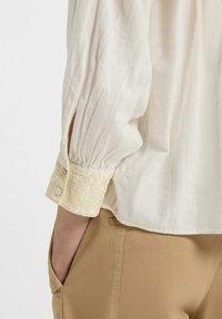 White textured blouse with puffed sleeves, featuring a decorative light yellow embroidered cuff. Paired with beige trousers.