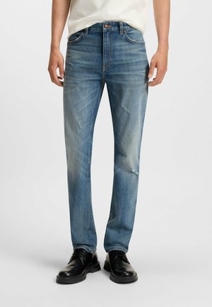 Light blue denim jeans with a straight cut, featuring fading and distressed details. Worn with black lace-up shoes.