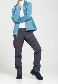 Blå fleecejacka, vit polotröja, grå cargobyxor och svarta vandringsskor med teal-accenter. Materialet ser mjukt ut med en texturerad look.