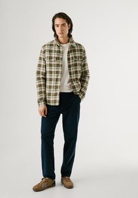 Chaqueta de franela a cuadros verdes con dos bolsillos en el pecho, camiseta de cuello redondo blanca, pantalones azul marino y zapatos marrones con acentos blancos.