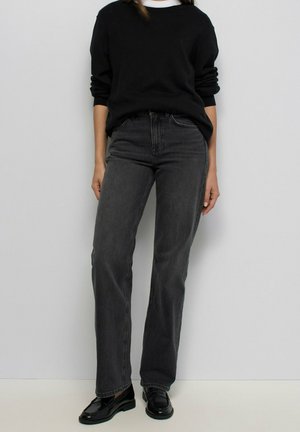 Persona con suéter negro de manga larga, jeans rectos de color gris oscuro y mocasines negros, de pie frente a un fondo gris claro.