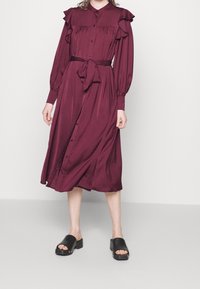 Burgundské dlouhé šaty s volánkovými rameny, zapínání na knoflíky a pásek v pase; nošeny s černými platformovými sandály.