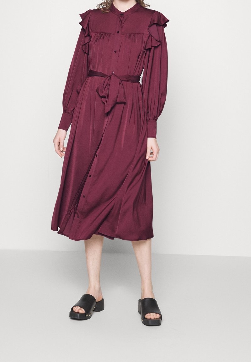 Burgundské dlouhé šaty s volánkovými rameny, zapínání na knoflíky a pásek v pase; nošeny s černými platformovými sandály.