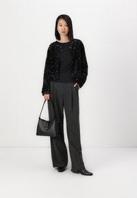 Giacca nera con paillettes, top nero, pantaloni grigio a vita alta, borsa nera, scarpe nere a punta; il modello è in piedi contro uno sfondo bianco.