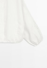 White long-sleeve blouse with elastic cuffs, lace trim along sleeves, a vertical button closure, and a small side pocket on front.