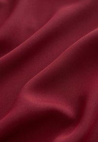 Burgundská tkanina s hladkým, mírně strukturovaným povrchem, která ukazuje jemné žebrování a vlnící se záhyby, zdůrazňuje její flexibilitu a splývavost.