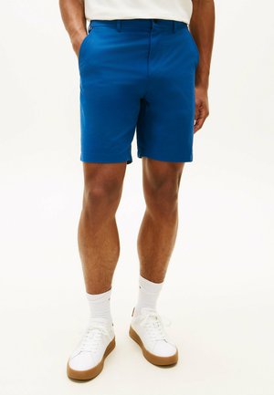 Hombre con pantalones cortos azules hasta la rodilla, calcetas blancas y zapatillas blancas con suelas de goma, de pie con las manos en los bolsillos.