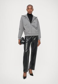 Szara polarowa bluza na zamek z haftowanymi detalami, zestawiona z błyszczącymi czarnymi spodniami z ekoskóry i czarnymi szpilkami. Modelka trzyma aparat.