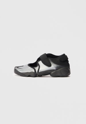 Scarpa atletica nera e argentata con un design traspirante, cinturino in velcro, punta arrotondata e suola testurizzata, caratterizzata da uno stile minimalista.