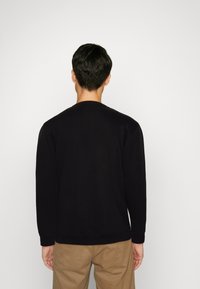 Černá mikina s dlouhým rukávem a kulatým výstřihem, s ribbenými manžetami a lemem. Hladký povrch, minimalistický design, nošená s béžovými kalhotami.