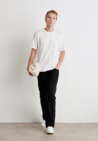 Hvit bomull t-skjorte, løs passform, med brodert logo. Sorte bukser med rett snitt. Lyst farget caps holdt i den ene hånden. Hvit sneakers.