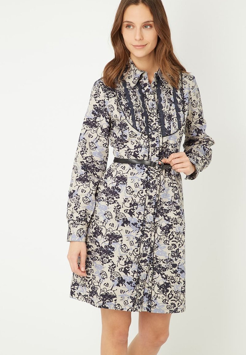 Vestido de manga larga en color crema con un patrón floral en azul y navy. Presenta un cuello con volantes y un cinturón en la cintura. Tejido suave con un brillo moderado.