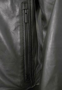 Schwarze Lederjacke mit einer Nahaufnahme des Reißverschlusses und der Nahtdetails. Das Leder hat eine glatte Textur mit subtiler Ausstrahlung und sichtbaren Nähten.