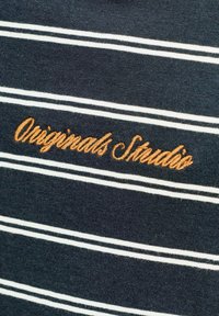 Tela azul oscuro con rayas horizontales blancas y texto bordado en naranja que dice "Originals Studio" en cursiva.