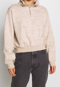 Beige-vetoketjullinen pusero, jossa on kevyt kuvio, pehmeä kangas, resorit hihansuissa ja helmassa, yhdistetty tummanharmaisiin farkkuihin.