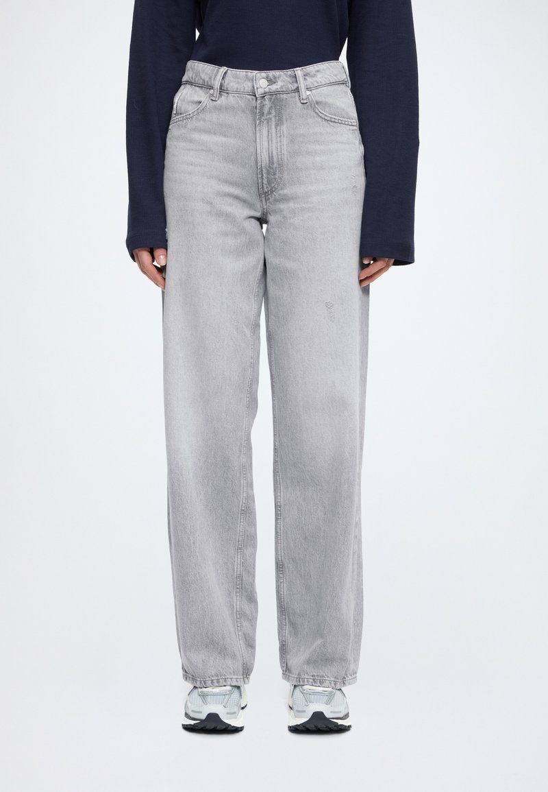 Sivé rifle s vysokým pásom a rovnými nohavicami, vyrobené z džínsoviny. Materiál má jemne opotrebovaný vzhľad. Kombinované s modrými teniskami a dlhým rukávom na vrchu.